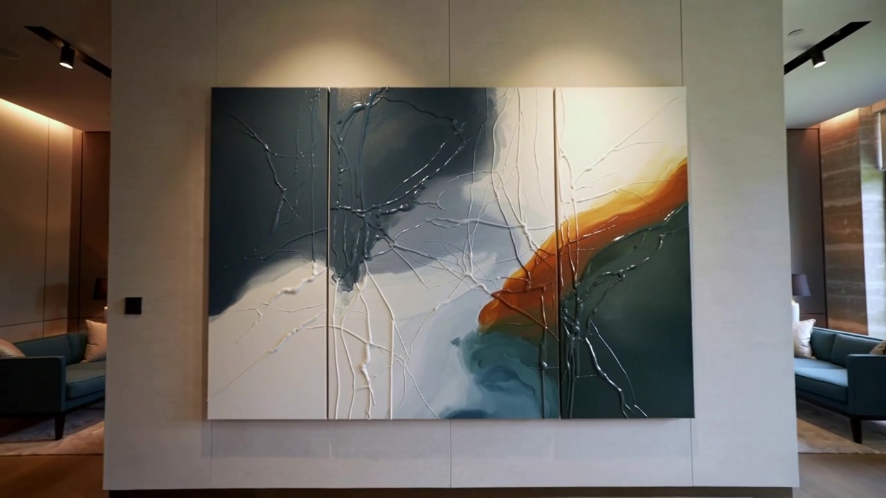 La telecamera si allontana da un primo piano, mostrando un trittico testurizzato sulla parete, rivelando l'opera d'arte completa e la stanza.