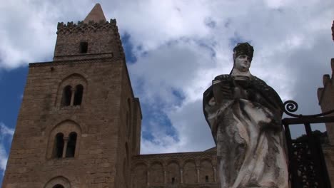 Ein-Wolkenzeitraffer,-Der-Sich-über-Einer-Statue-Einer-Frau-Bewegt,-Die-Ein-Buch-Und-Ein-Gebäude-In-Cefalu,-Italien-Hält