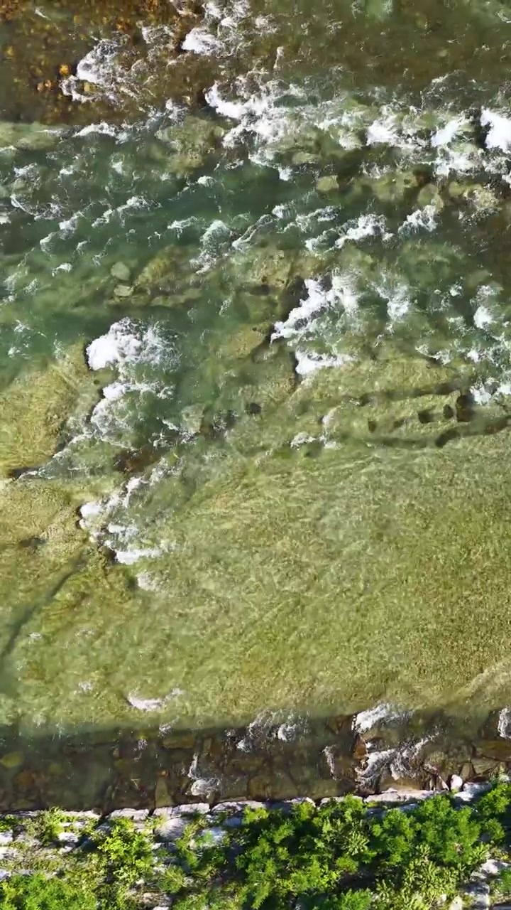 Ripresa aerea con drone dall'alto cattura acqua limpida del fiume che scorre impetuosa sulle pietre lungo una sponda verde.