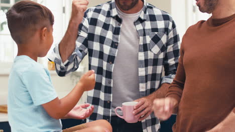 familia del mismo sexo con dos padres en la cocina con el hijo sentado en el mostrador jugando tijeras de papel de roca