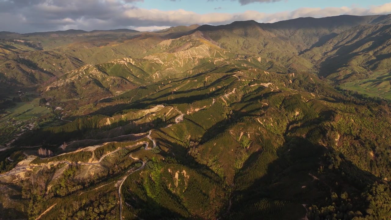 Cordilheira acidentada com trilhas de terra sinuosas esculpidas em encostas douradas, cristas em camadas e vales florestais banhados pela luz quente do nascer do sol sob nuvens dramáticas. Nova Zelândia. Tomada aérea ampla em retrocesso.