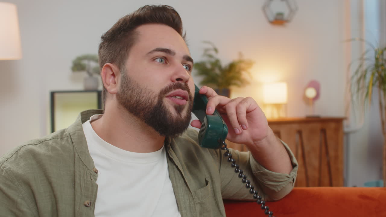 Smiling young man making wired telephone call conversation with friends sitting on sofa at home
