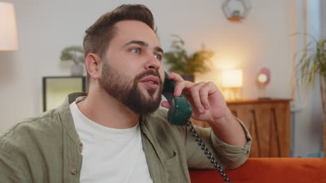 smiling young man making wired telephone call conversation with friends sitting on sofa at home