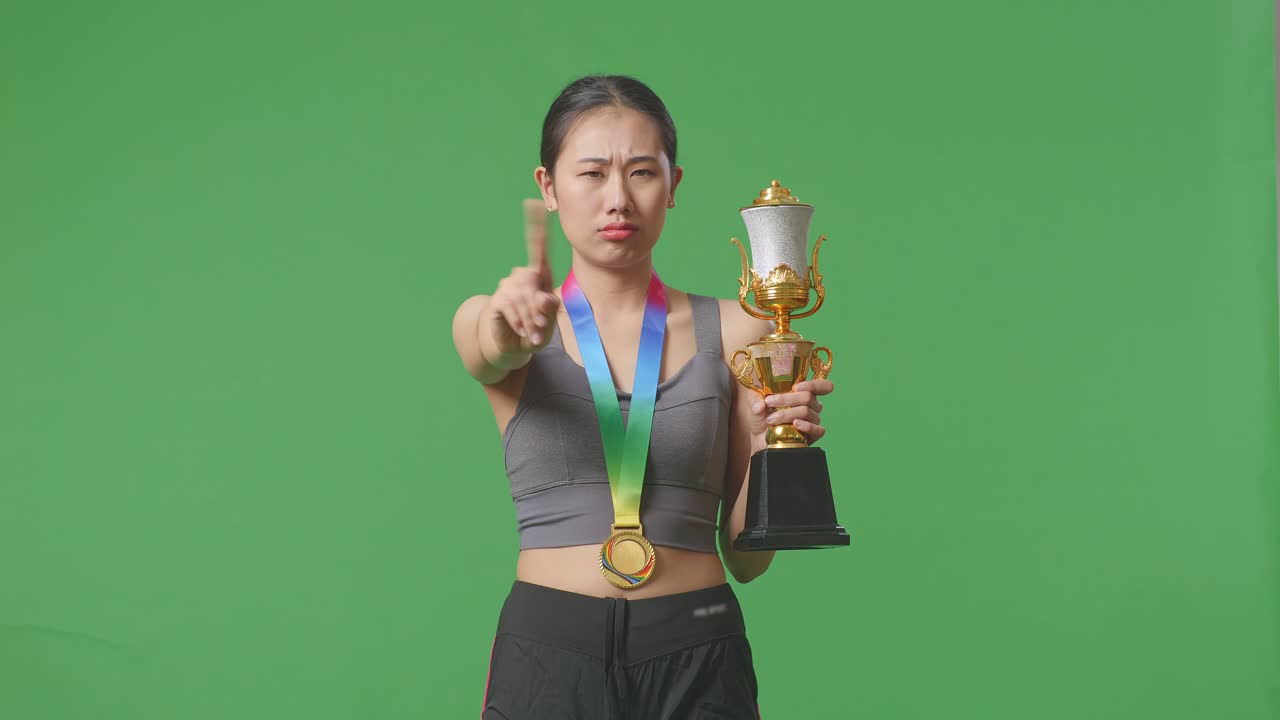 mujer asiática con una medalla de oro y trofeo desaprobando sin señal del dedo índice y sonriendo a la cámara en el fondo de la pantalla verde en el estudio