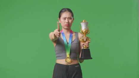 mujer asiática con una medalla de oro y trofeo desaprobando sin señal del dedo índice y sonriendo a la cámara en el fondo de la pantalla verde en el estudio
