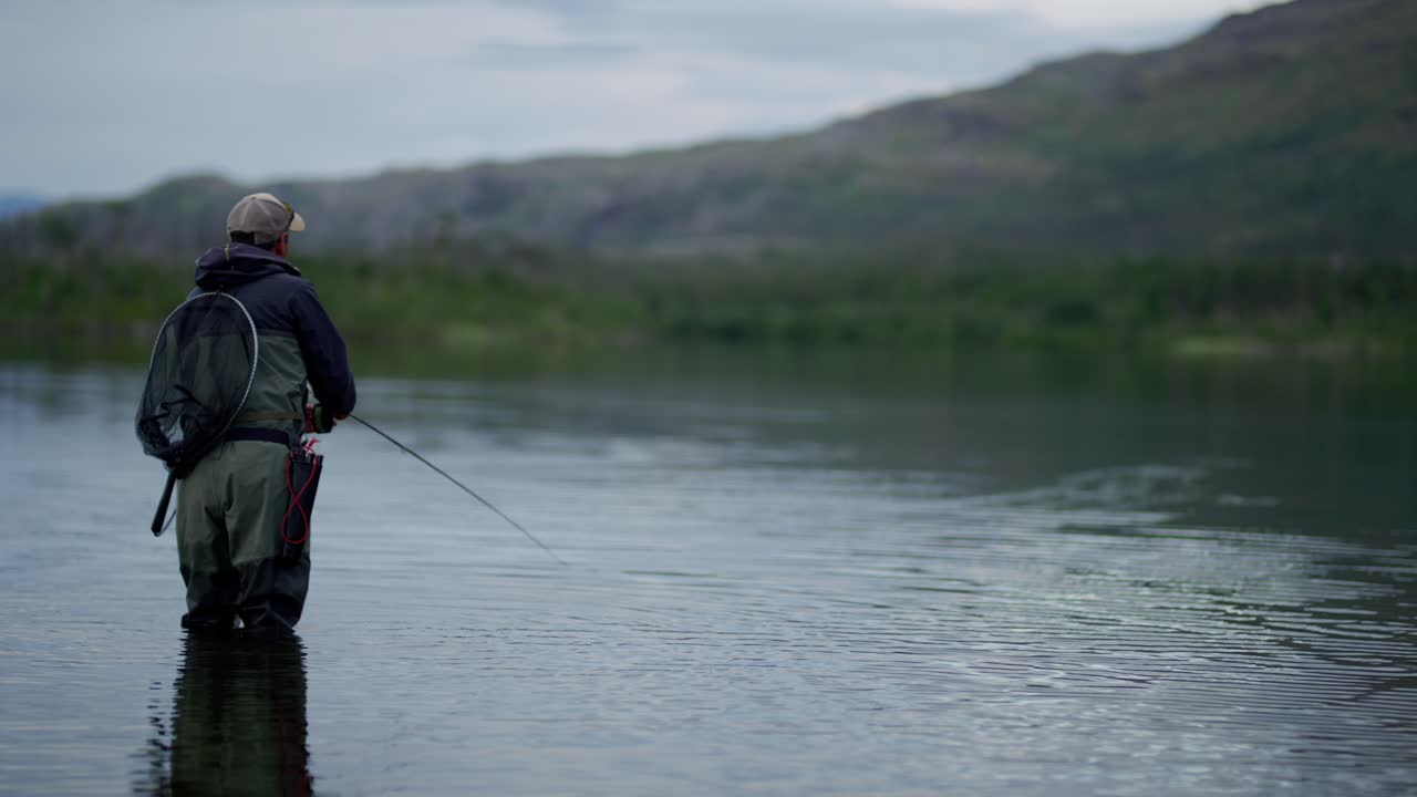 Man casts fly fishing rod in still Swedish lake, slomo from behind