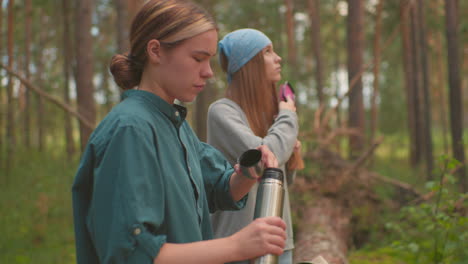 hikers take peaceful break in lush forest, one carefully opens thermos while other gently brushes hair, backpacks rest on fallen tree, capturing relaxed, serene moment as they enjoy nature