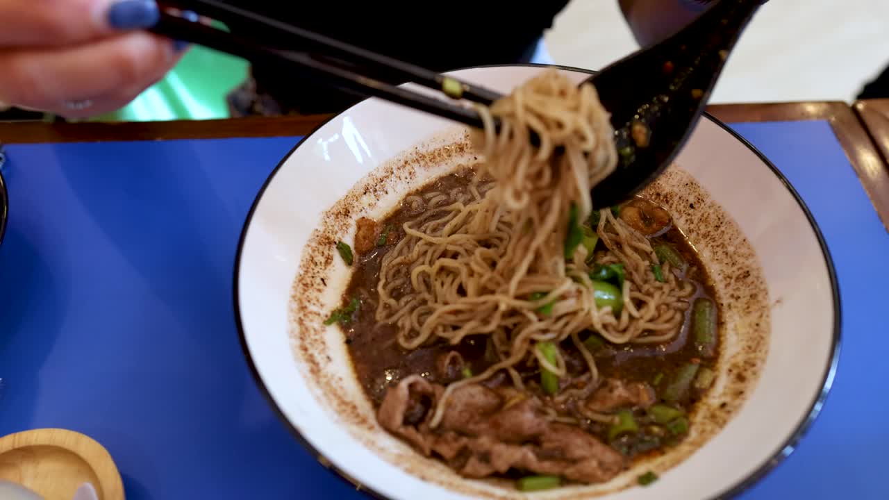 Primo piano di mani che usano le bacchette per sollevare vermicelli di riso da una zuppa di maiale scura.