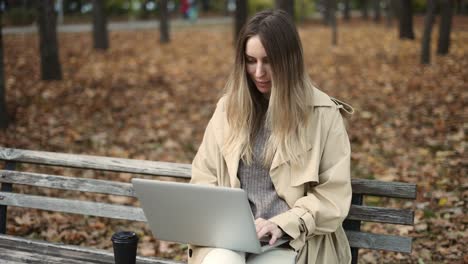 Young-beautiful-woman-works-at-a-laptop,-sitting-on-a-park-bench-with-coffee