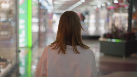 back view of a lady in a white top walking through a shopping mall and looking around, surrounded by colorful lights and a blurred background featuring glass elements