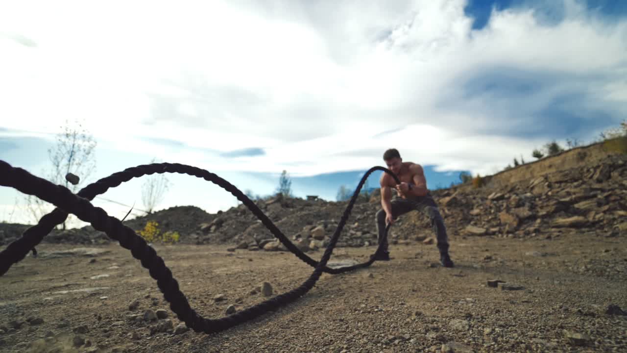 Sportsman exercising with battle ropes outdoors. Strong athlete doing double and alternating wave exercise with battle ropes standing among rocks in nature.