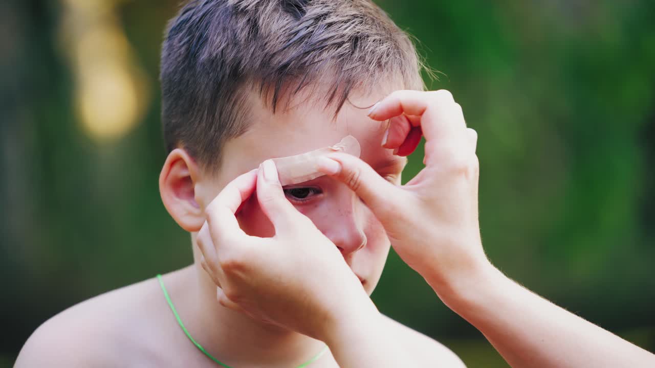Sad boy with injury on his eyebrow. Woman's hands putting small white adhesive plaster on a wound outdoors. Close-up.