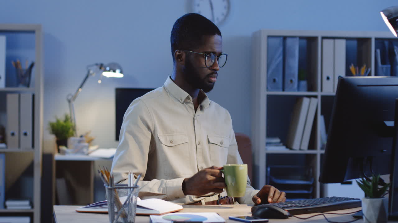 Young Man Drinking Coffee While Working At The Computer In The Office At Night