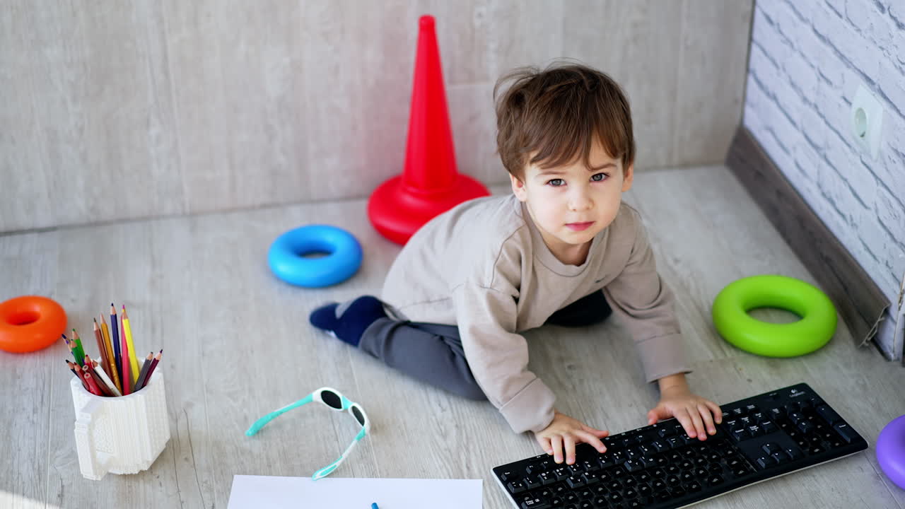 Dark-haired toddler sitting on the floor busy with computer keyboard. Baby boy playing at home. Top view.