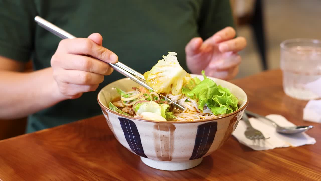 Lähikuva henkilöstä syömässä vietnamilaista bun chaa metallisilla syömäpuikoilla ravintolassa.
