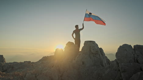 Drone-shot-of-a-hiker-standing-on-top-of-mountain-Krn-with-a-slovenina-flag-attached-on-a-hiking-pole-in-early-morning-hours,-clear-sky,-sun-shining
