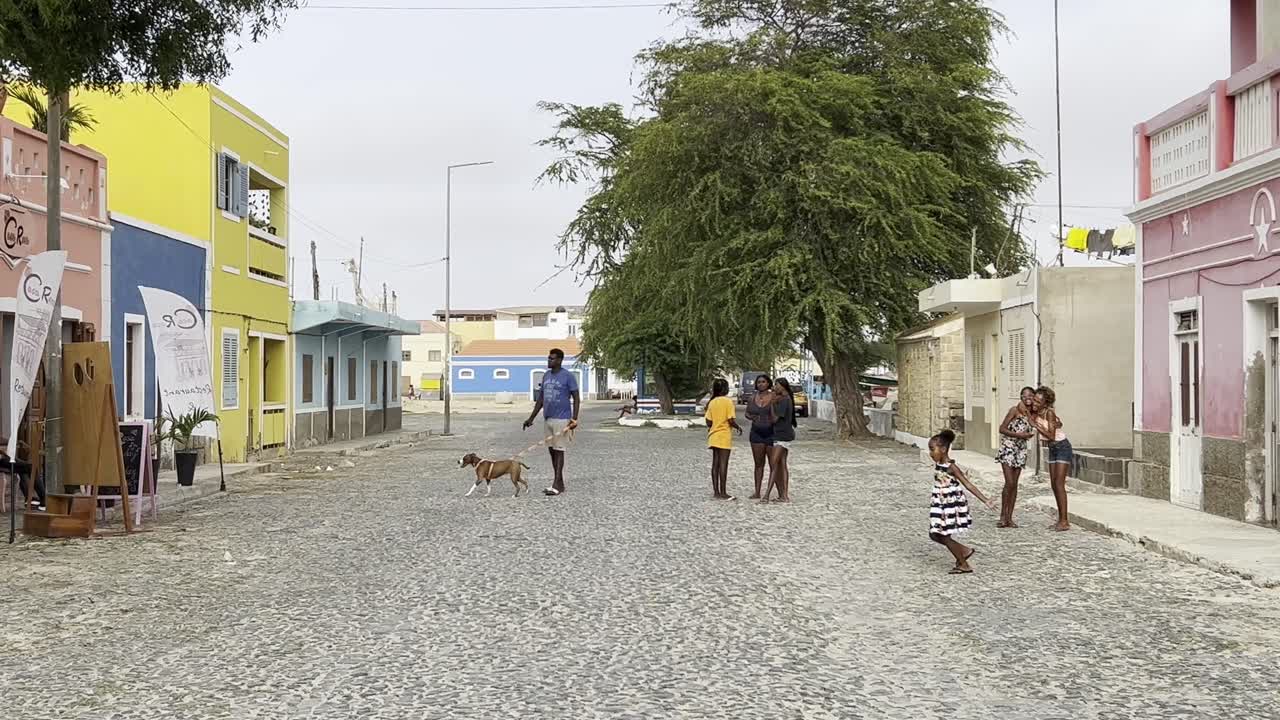 Tranquil cobbled street in Sal Rei, Cabo Verde, lined with colorful colonial-style buildings, trees, and locals enjoying the day. The warm, inviting scene captures the essence of island life