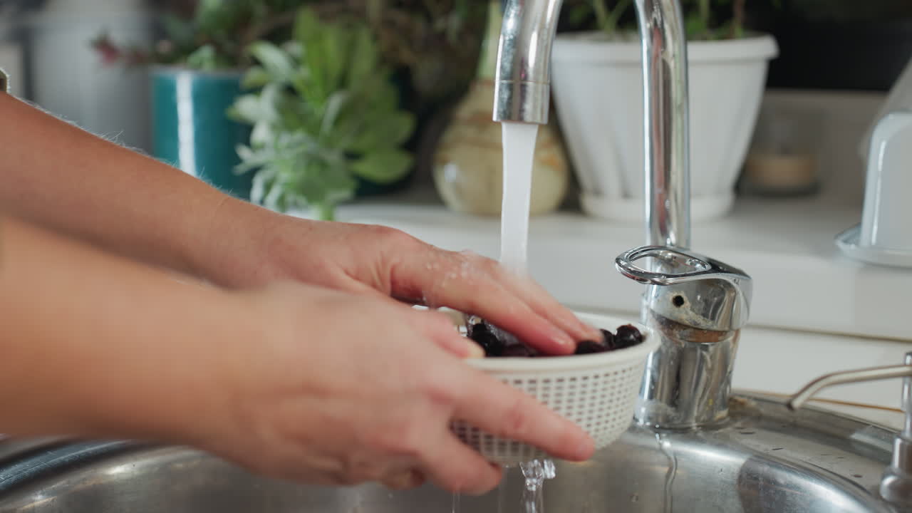 Close up hand view of person turning on kitchen tap to rinse fresh dark berries in white strainer bowl, preparing for washing, with potted plants in background and stainless sink under tap stream
