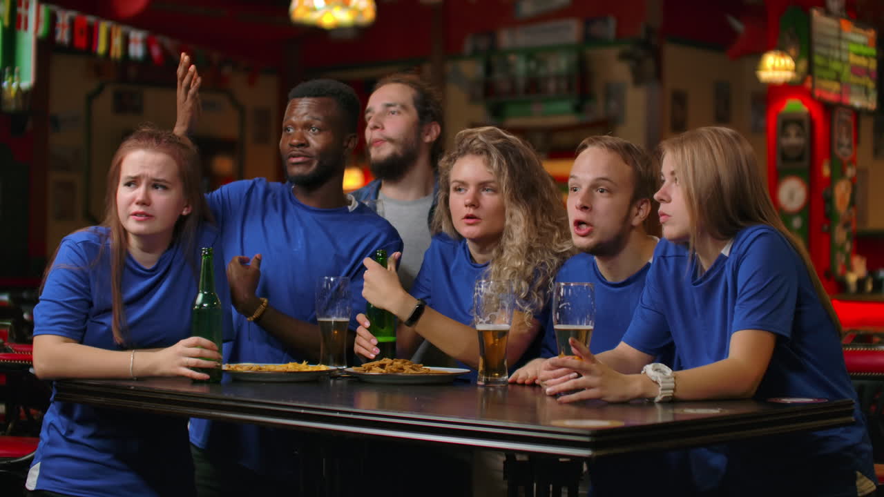 un grupo de amigos de los aficionados en camisetas azules emocionadamente emocionalmente ver el partido y regocijarse en el gol anotado saltar y gritar emocionalmente. celebrar y celebrar un gol anotado masticando con cerveza.