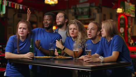 A-group-of-friends-of-fans-in-blue-t-shirts-excitedly-emotionally-watch-the-match-and-rejoice-at-the-goal-scored-jump-and-shout-emotionally.-Celebrate-and-celebrate-a-goal-scored-by-chewing-with-beer.