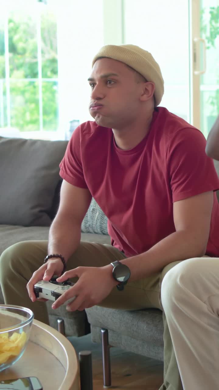 Video verticale: Gioco che si avvia, uomo con maglietta rossa e cappello sul divano che tiene un controller bianco e fa uno spuntino.