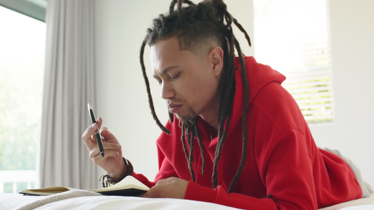 Window inspiring mid adult man lying on bed, flipping pages, writing, checking pen, smiling