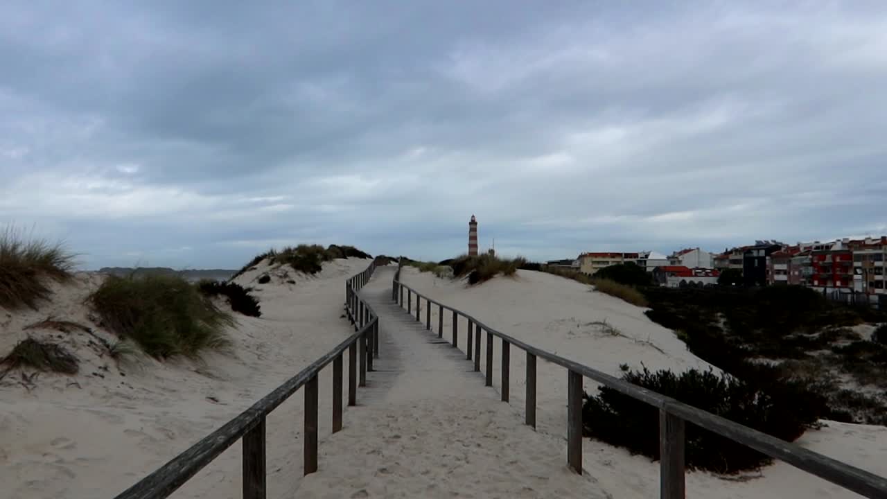 un sendero de madera arenosa conduce a la playa de costa nova con el faro de barra, cielo nublado sobre la duna costera