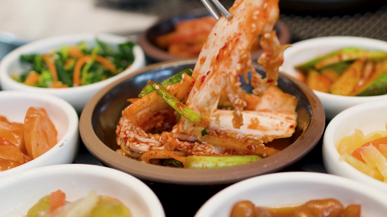 Tampilan close-up sumpit logam mengangkat kubis fermentasi pedas (kimchi) dari berbagai lauk pauk.
