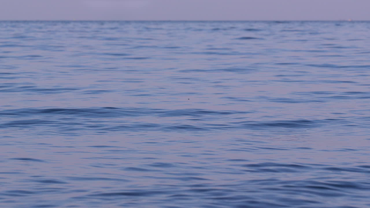 Smooth blue water under pastel evening sky, subtle waves forming meditative looping texture for backgrounds