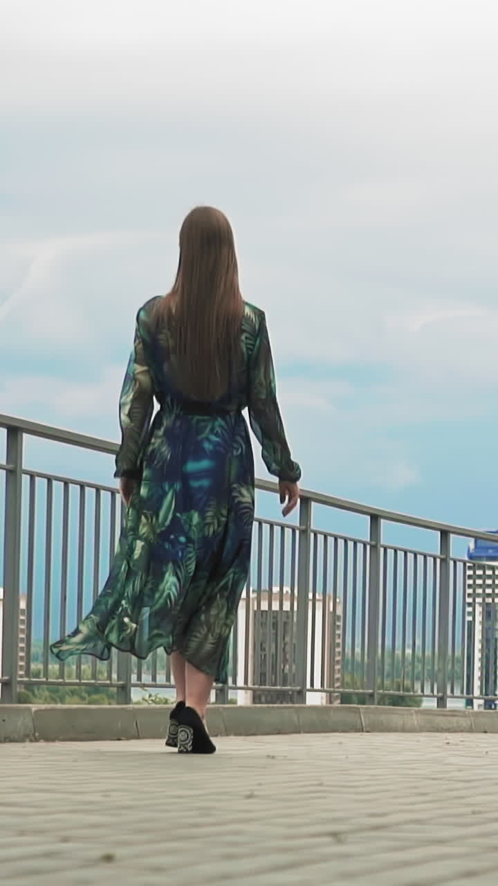 Young slim woman in stylish long dress and shoes with high heels walks along viewing deck in contemporary city slow motion backside view