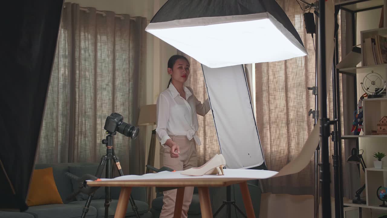 fotógrafa asiática ajustando el equipo de iluminación para obtener una buena iluminación en los zapatos de las mujeres mientras toma fotos de los productos en el estudio doméstico