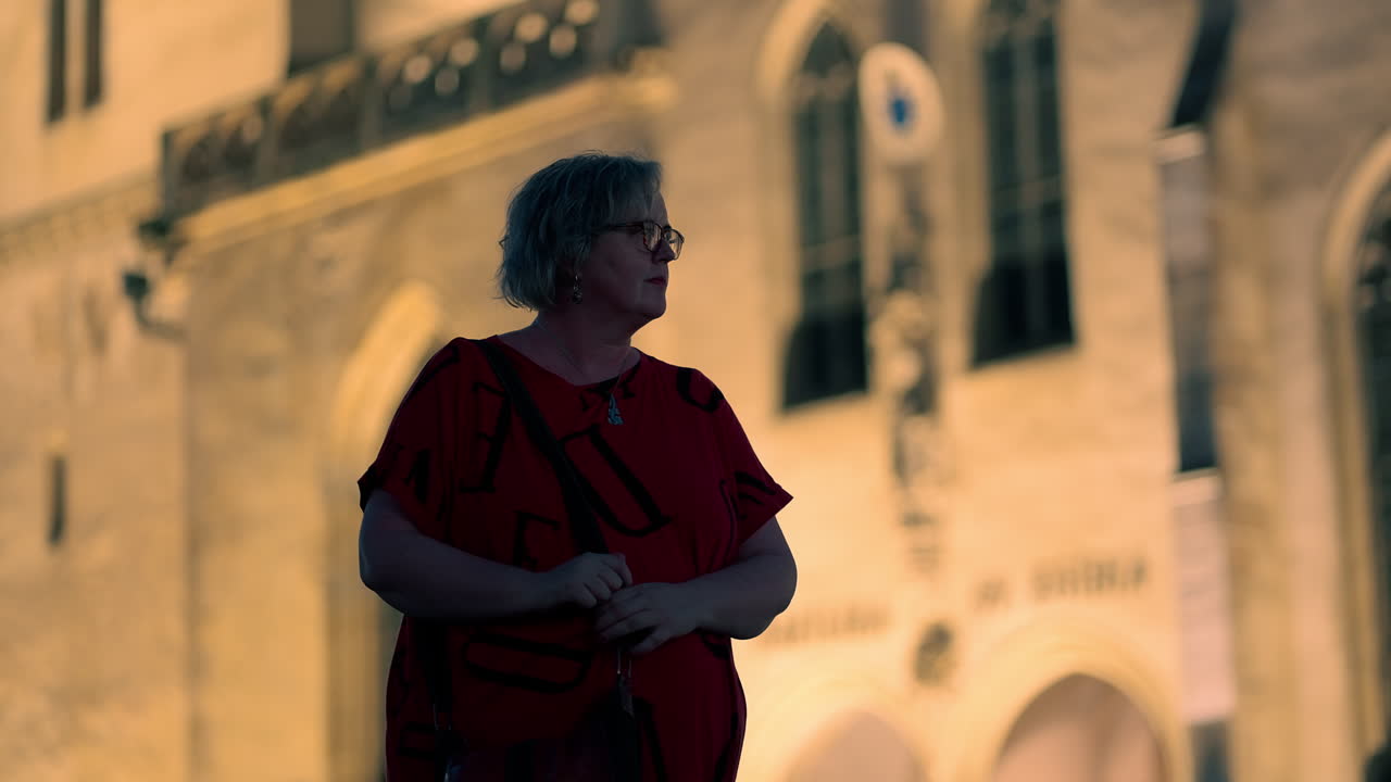 Donna anziana bianca con gli occhiali in piedi e che si guarda intorno davanti a una chiesa storica europea illuminata di notte al rallentatore.
