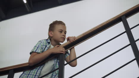 Curious-schoolboy-standing-stairway-alone.-Boy-leaning-on-railings-examine-hall.