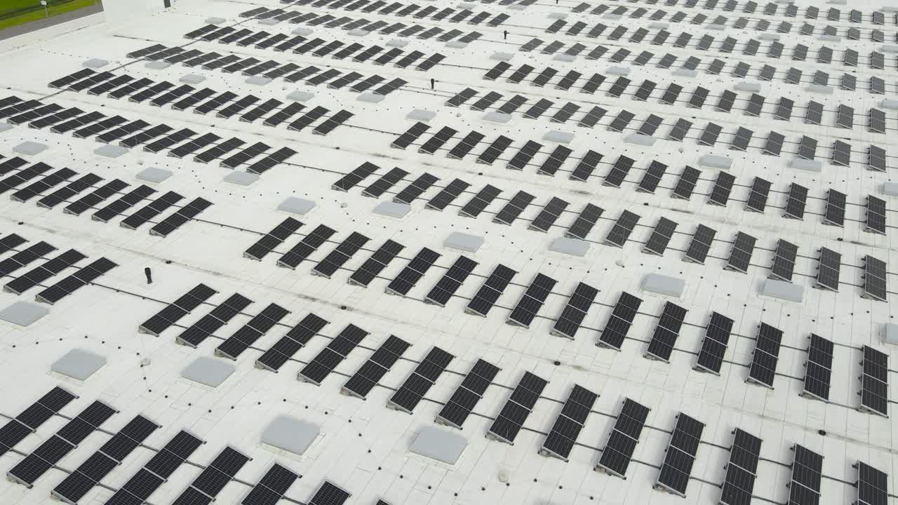 Gorgeous aerial flyover view of hundreds of green and sustainable energy solar panels on a rooftop collecting sun and day light. The black panels are next to each other with equal spaces on white roof