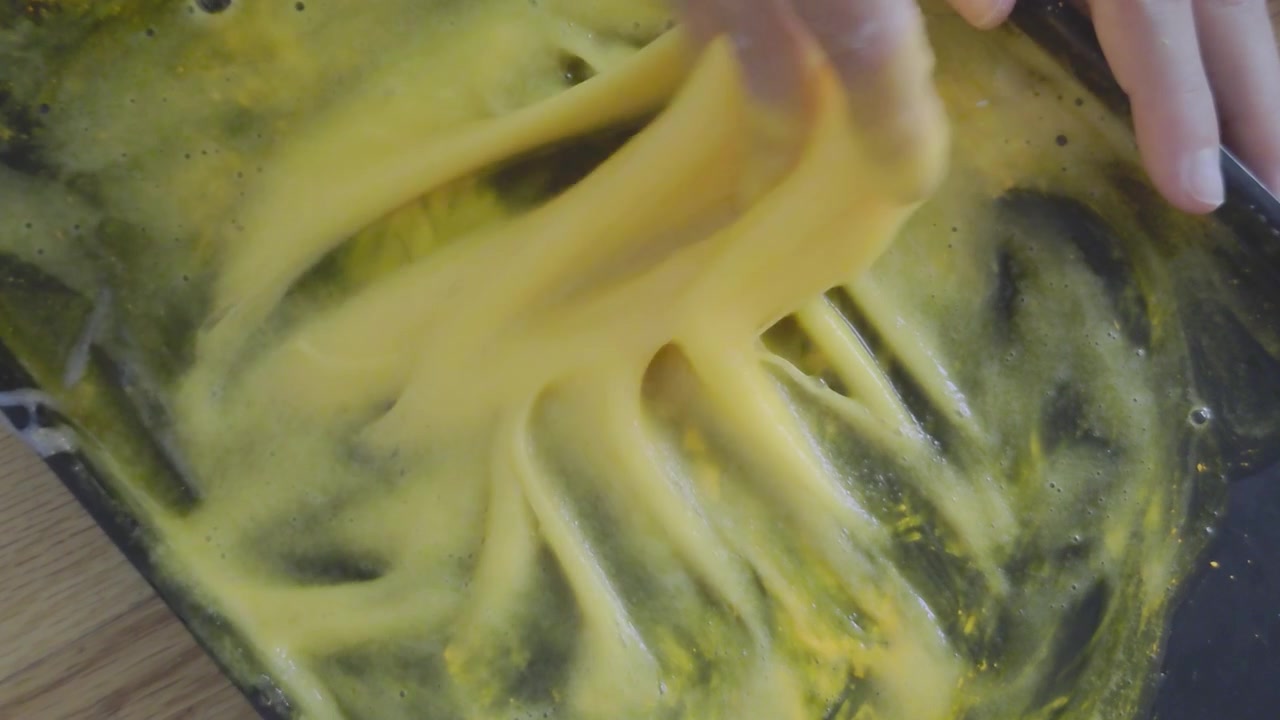 A high angle close-up shot captures hands actively kneading, mixing, and pressing down on thick, sticky yellow slime inside a dark container during a popular home science experiment and tactile STEM c