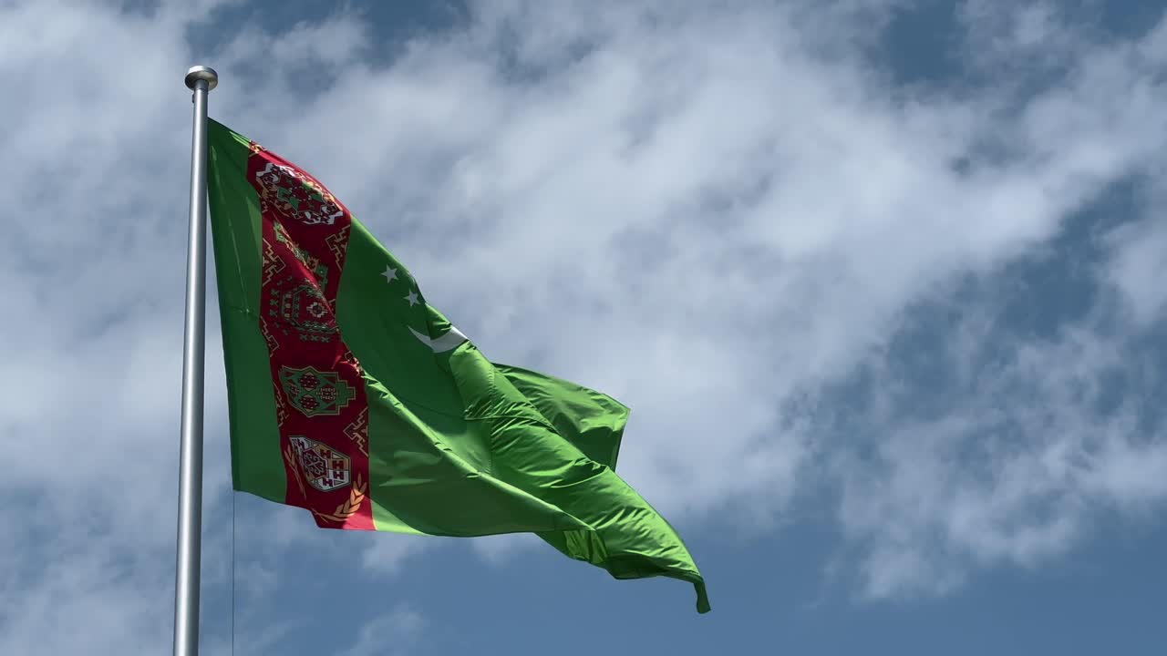 A close-up shot of the flag of Turkmenistan fluttering vibrantly in the wind under a bright sky, showcasing its rich green color and intricate carpet pattern in stunning detail