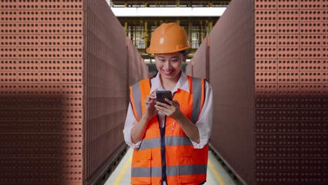 asian female engineer with safety helmet using smartphone while standing with red brick packed in stacks are stored
