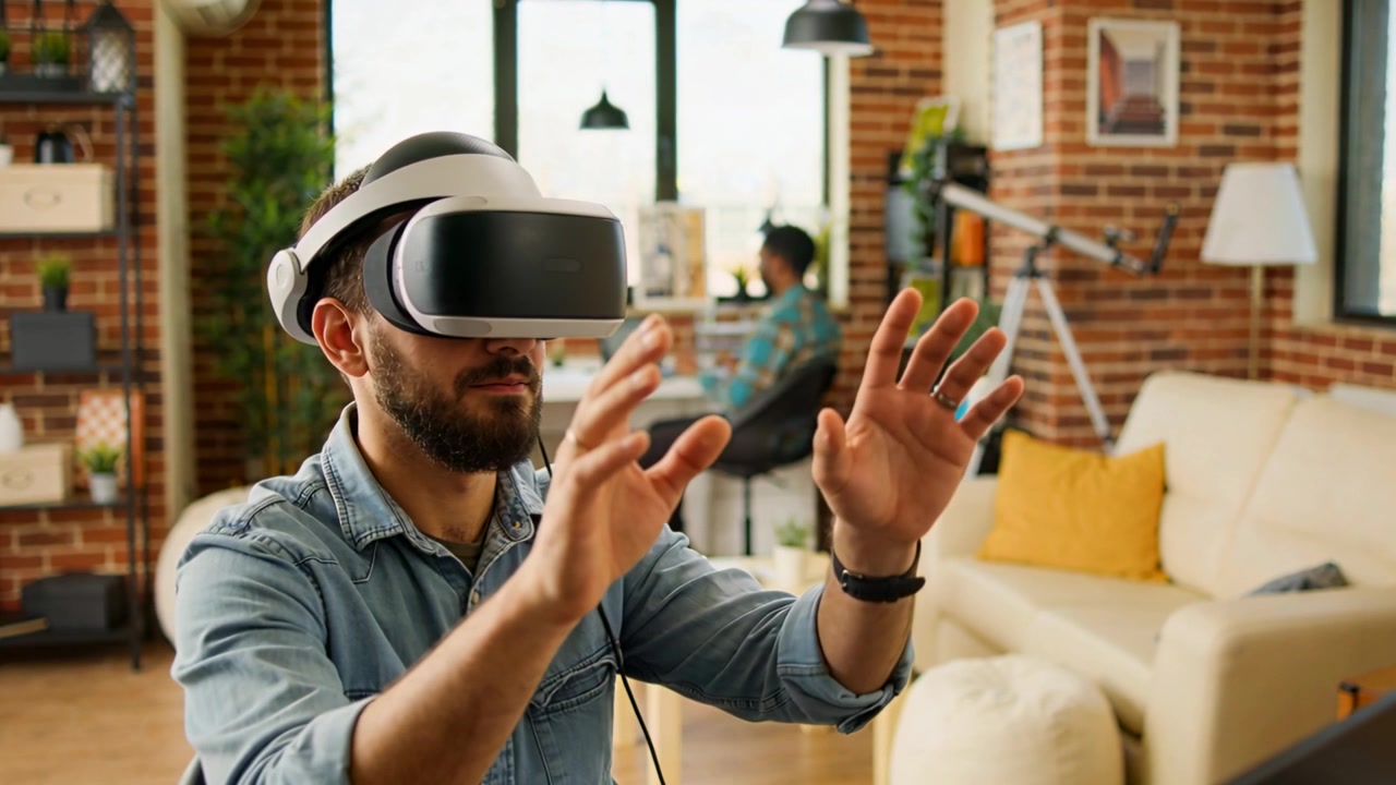 Uomo in camicia di jeans con visore VR che si protende per afferrare oggetti virtuali in un loft, laptop sullo sfondo.