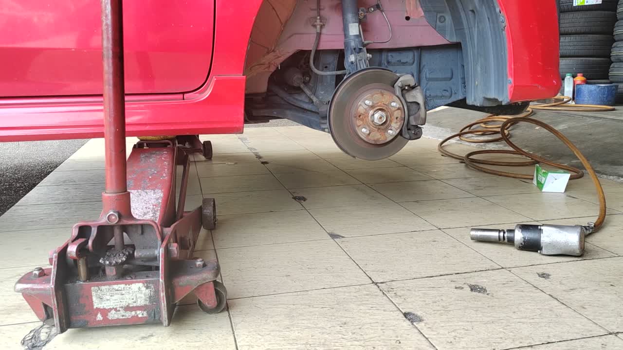 A small red car is seen in a workshop, undergoing a tire change. The front tire is replaced as the car is slightly lifted for ease.