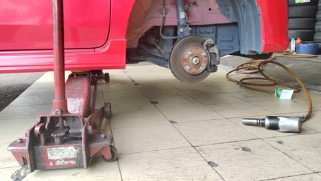 a small red car is seen in a workshop, undergoing a tire change. the front tire is replaced as the car is slightly lifted for ease.