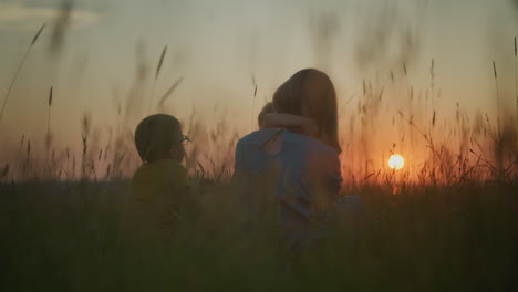 a back shot of a mother in a blue gown sitting in a field during sunset, holding her younger child on her lap who embraces her neck. the older child sits beside them as they all gaze out at the sunset