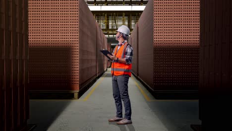 full body side view of asian male engineer with safety helmet taking note on the tablet and looking around while standing with red brick packed in stacks are stored