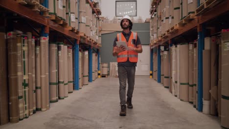 a young male worker maintaining records using digital tablet waking through the large warehouse with freight stacked