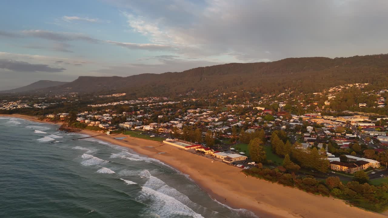 Coastal town of Thirroul stretches beside ocean with rolling surf and residential homes, aerial angled approach