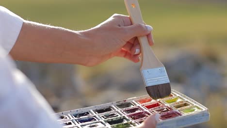 close-up of a hand dipping a paintbrush into a watercolor palette