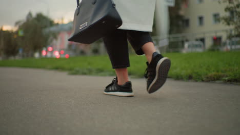 partial view of girl holding designer handbag and twirling closed umbrella while walking along paved path with blurred background featuring green grass, office building, and passing vehicles