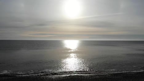 aerial flying out towards calm sea waves with sun being reflected on surface