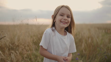 a happy smiling little girl looking at the camera runs in slow motion at sunset in a field of grain ears