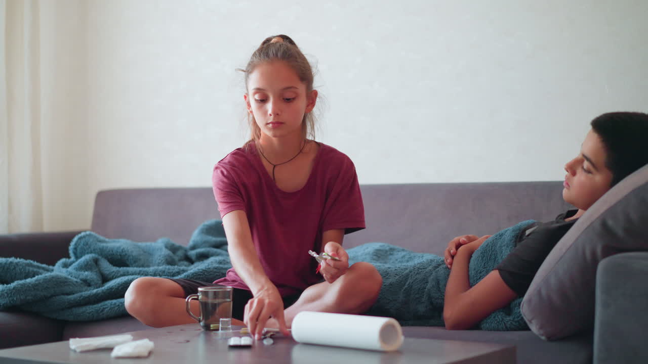 Young girl prepares medicine in cup using spoon while caring for sick brother lying on couch under blanket in cozy home setting with calm and caring atmosphere during illness recovery