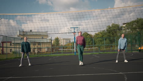 volleyball players standing in court with lady in green leggings preparing to slam volleyball, background features volleyball net and building in an outdoor sports arena